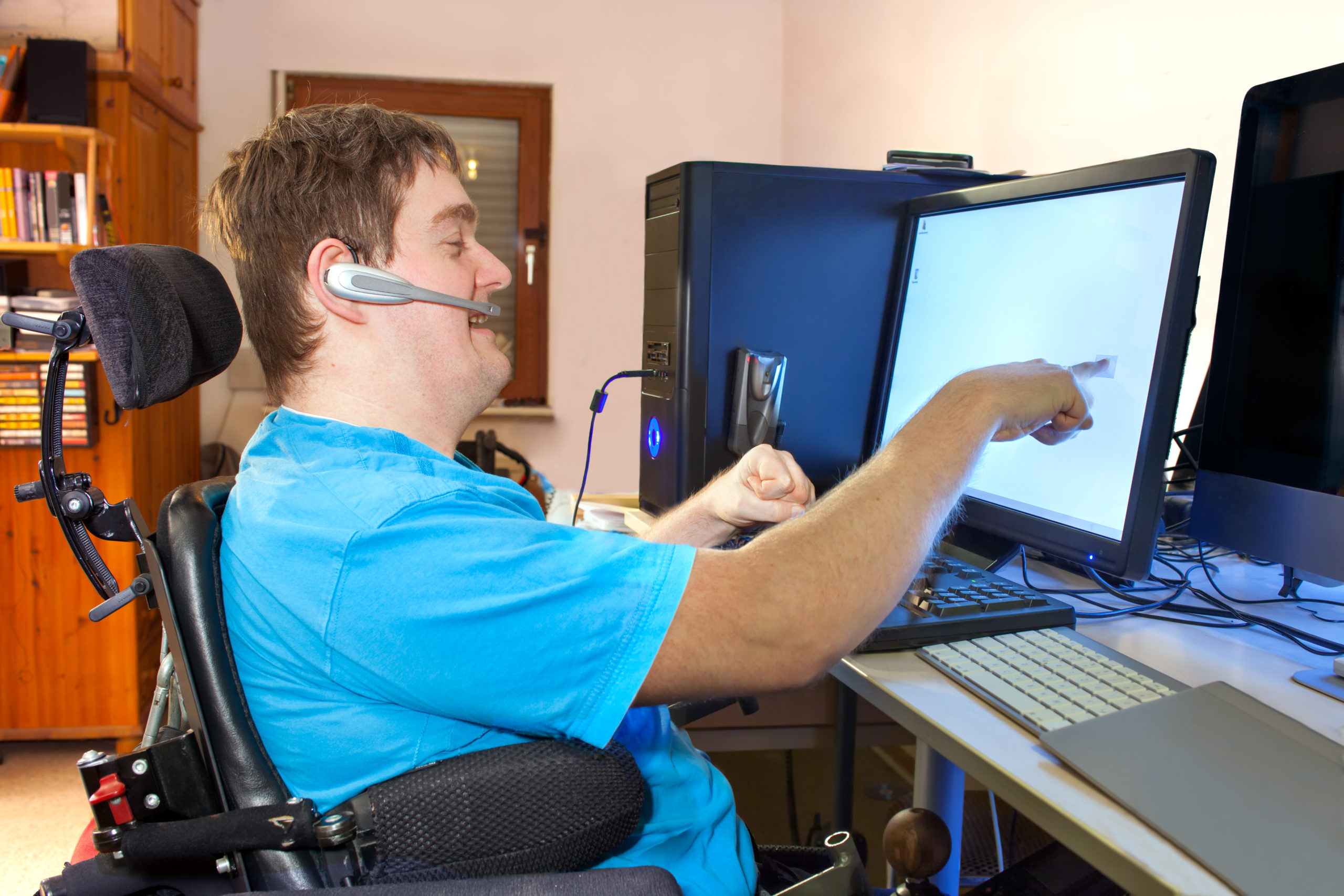 Image of a man with cerebral palsy in a wheelchair using adaptive technology on a computer. The man is wearing a blue tshirt and a bluetooth headset, and is smiling